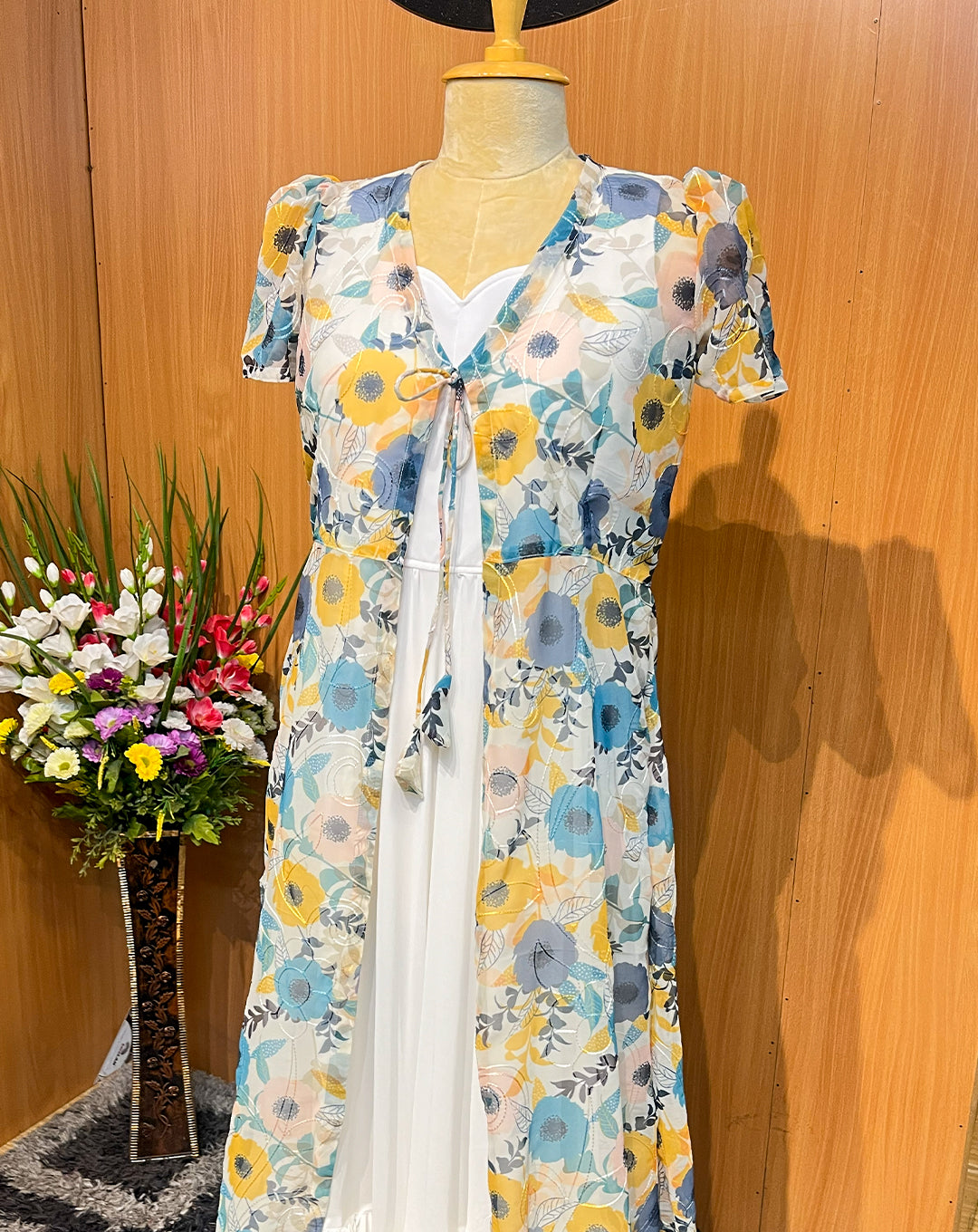 This elegant sleeveless white georgette maxi is paired with a lightweight sheer floral overcoat, creating a soft and flowy silhouette perfect for brunches, parties, and holiday outings. The pastel flower print, tie-up front detail, and matching hair bow add a feminine, youthful charm, while the layered hemline gives graceful movement with every step.