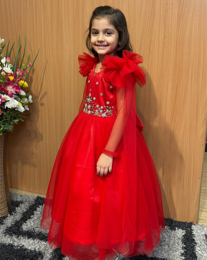 Young girl in a red dress with floral decorations standing against a wooden wall.