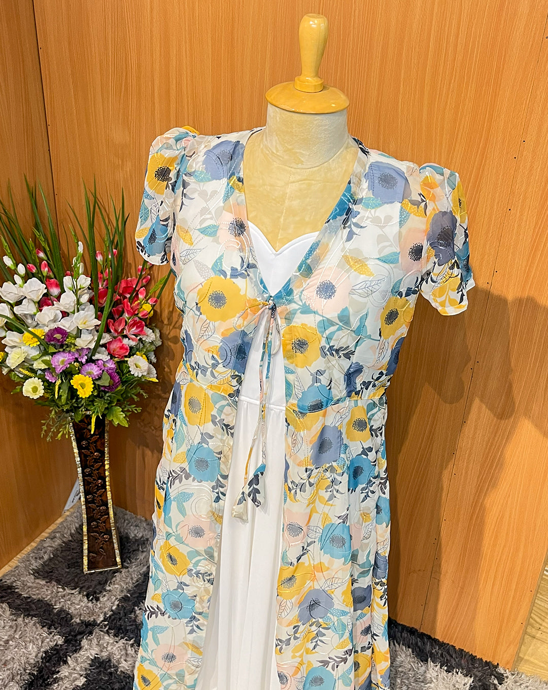 This elegant sleeveless white georgette maxi is paired with a lightweight sheer floral overcoat, creating a soft and flowy silhouette perfect for brunches, parties, and holiday outings. The pastel flower print, tie-up front detail, and matching hair bow add a feminine, youthful charm, while the layered hemline gives graceful movement with every step.