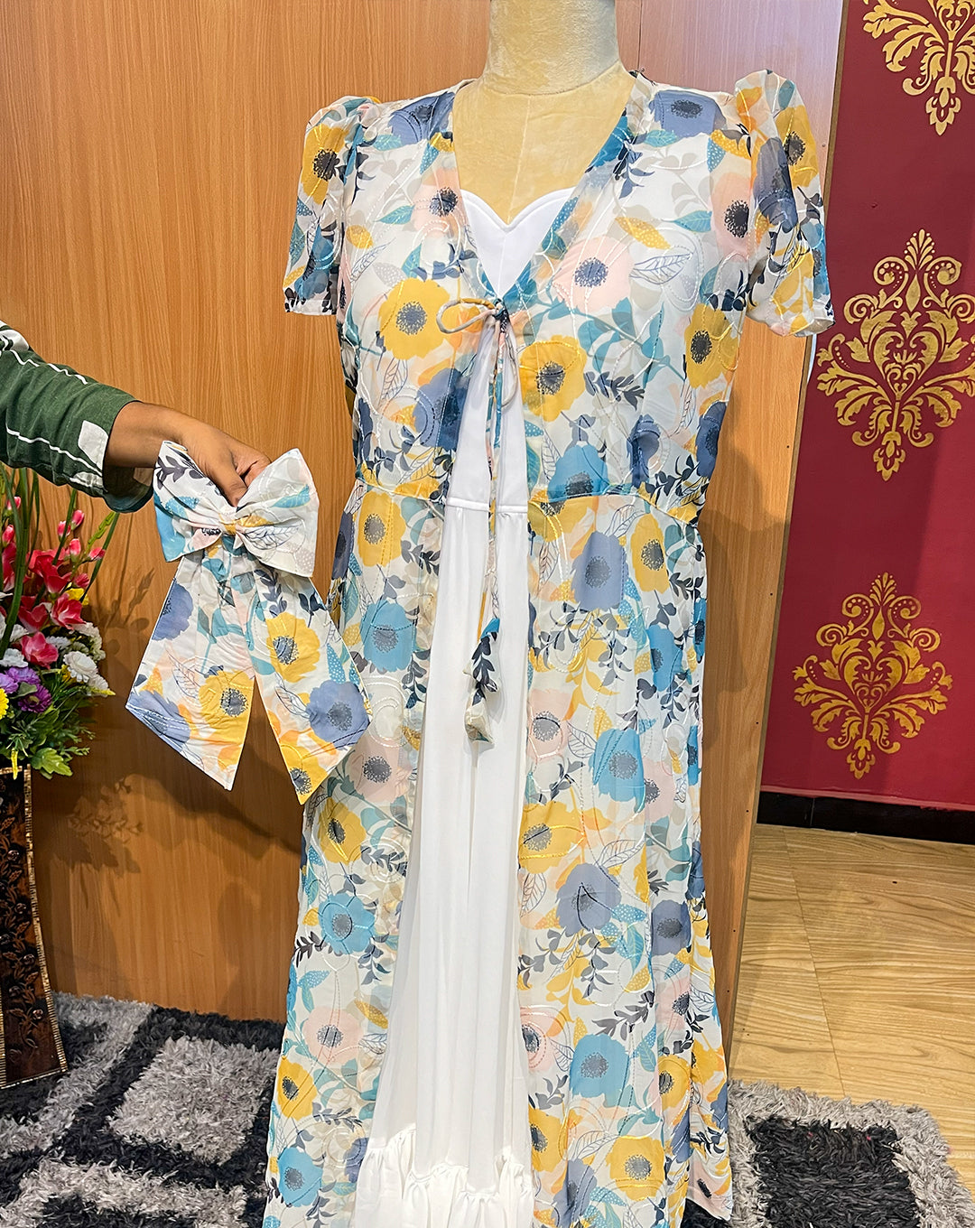This elegant sleeveless white georgette maxi is paired with a lightweight sheer floral overcoat, creating a soft and flowy silhouette perfect for brunches, parties, and holiday outings. The pastel flower print, tie-up front detail, and matching hair bow add a feminine, youthful charm, while the layered hemline gives graceful movement with every step.